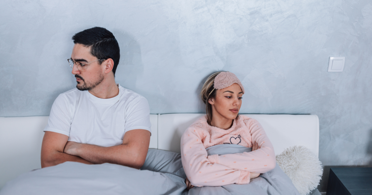 Ein Paar sitzt im Bett voneinander abgewandt und wirkt angespannt und emotional distanziert.