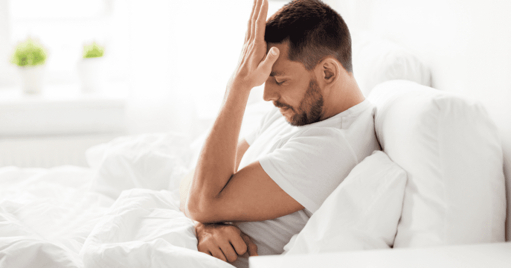 Mann sitzt nachdenklich im Bett und hält die Hand an die Stirn · Stress und Leistungsdruck bei erektiler Dysfunktion