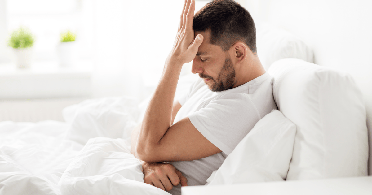 Mann sitzt nachdenklich im Bett und hält die Hand an die Stirn · Stress und Leistungsdruck bei erektiler Dysfunktion