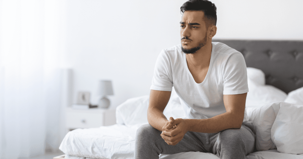 Erektile Dysfunktion Ursachen: Mann sitzt nachdenklich auf dem Bett · Stress, Unsicherheit und Leistungsdruck
