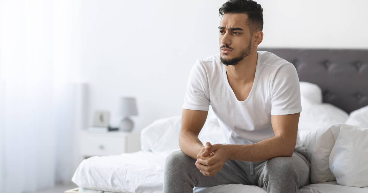 Erektile Dysfunktion Ursachen: Mann sitzt nachdenklich auf dem Bett · Stress, Unsicherheit und Leistungsdruck
