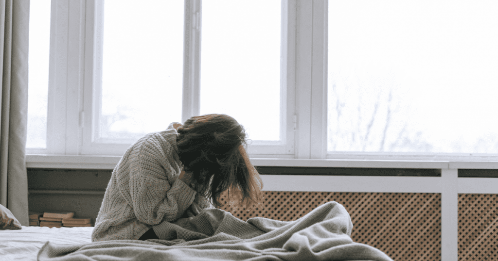 Vaginismus Behandlung: Frau sitzt angespannt im Bett und hält den Kopf · Stress, Scham und Rückzug bei Schmerzen