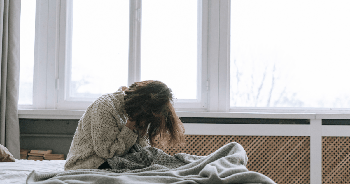 Vaginismus Behandlung: Frau sitzt angespannt im Bett und hält den Kopf · Stress, Scham und Rückzug bei Schmerzen