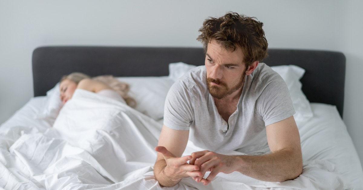 Mann sitzt nachdenklich im Bett und schaut auf sein Handy – passend zum Thema morgendliche Erektion, Leistungsdruck und Erektionsprobleme beim Sex.